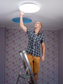 A man with a stepladder sets the ceiling lamp in the room.