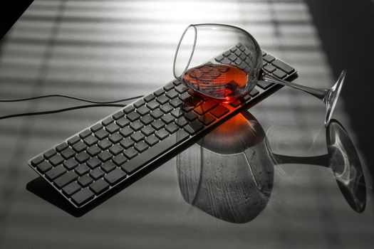 A glass of red wine lies on the keyboard on a white table with a shade from the blinds.