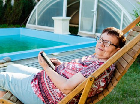 An elderly retired woman sitting by the pool in the backyard of the house with interest reading an e-book.