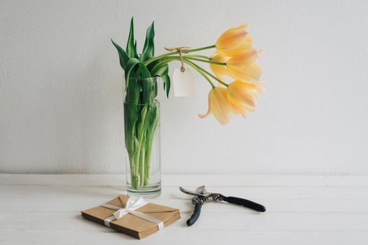 The secant-cut fragrant tulips stand in a glass vase. The tag is tied to a bouquet of yellow tulips. The envelopes of kraft paper are tied with a white satin ribbon. White wooden table.