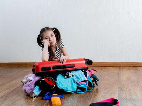 Sad girl leaned on a red suitcase that does not close because of a large number of things. Opening of borders. A little girl is going on vacation