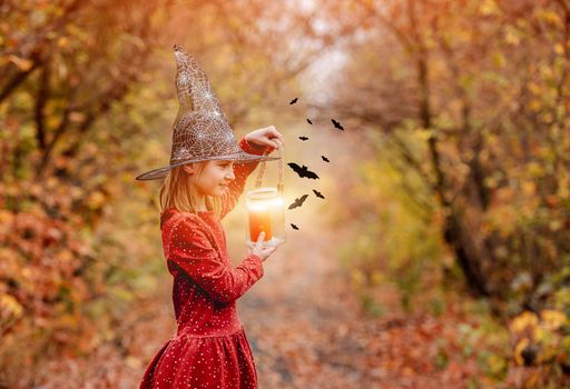Little girl in witch hat with candle attracting bats standing sideways in bright autumn wood