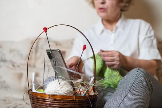 Faceless old woman knits and watches online training on a smartphone. Close-up of female hands with yarn and knitting needles