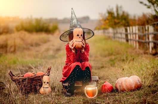 Little girl in halloween costume sitting next to pumpkins while frightening at camera on autumn nature