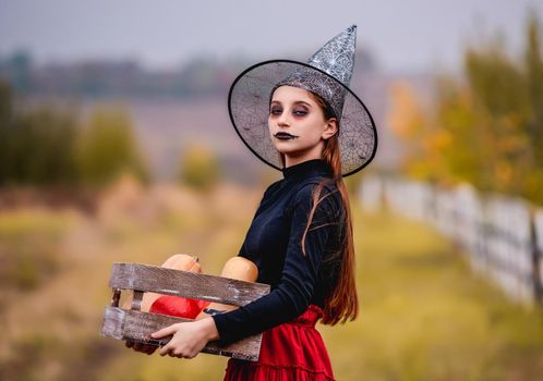 Gothic young girl on halloween in witch costume holding pumpkins box on nature background