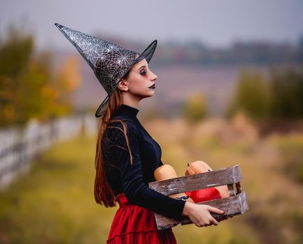 Gothic young girl on halloween in witch costume holding pumpkins box on nature background