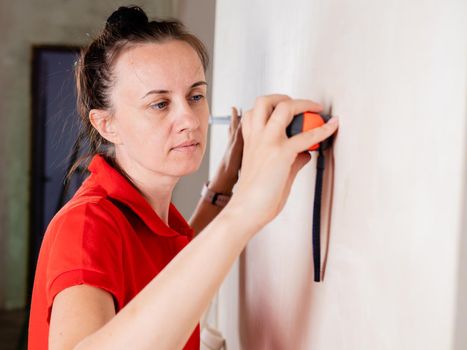 A woman uses a measuring tape measure to measure the wall for wallpapering.