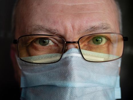 Close-up face portrait of senior man in medical mask and glasses looking at camera indoors.