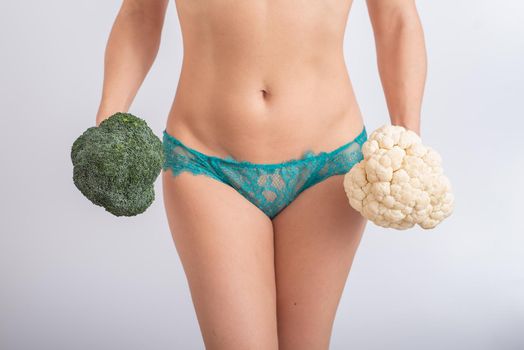 A faceless woman in panties holds cauliflower and broccoli on a white background. Food habits