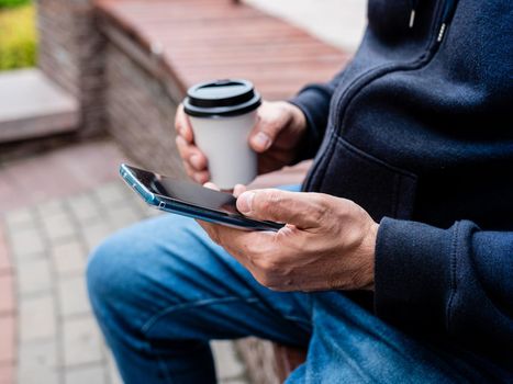 A man sits on a bench drinking coffee from a paper Cup and use a smartphone, close-up.