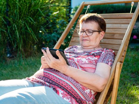 An elderly woman is sitting on a wooden deck chair in the backyard of a house and reading an e-book. Spending time in retirement.