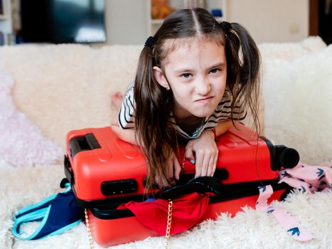 A little girl has Packed too many things on vacation, and can't close the red suitcase.
