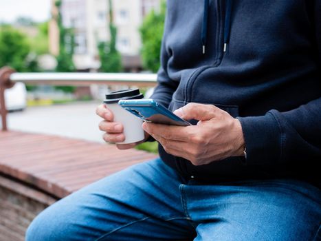 A man sits on a bench drinking coffee from a paper Cup and use a smartphone, close-up.