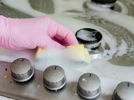 A woman in pink gloves washes the gas stove with a washcloth and detergent.