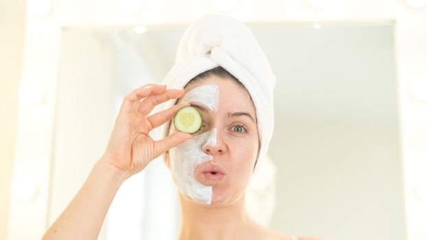Cheerful woman with a towel on her hair and in a clay face mask fooling around with cucumbers in her hands. Taking care of beauty at home.