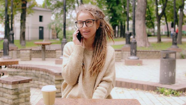 cheerful young caucasian woman with dreadlocks has phone conversation by phone in city park using smartphone