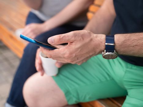 A man and a woman are sitting together on a bench in a street cafe, drinking coffee and using a smartphone. Close-up of a man's hands with a smartphone.