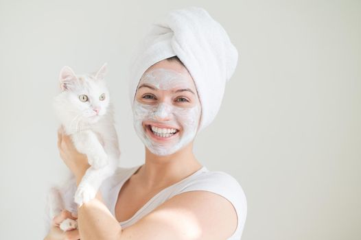 A woman with a towel on her hair and a clay mask on her face is holding a white fluffy cat on a white background. Copy space.