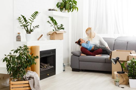 Cute mother and little daughter are on floor near boxes. They are preparing to move in another building. The parent and child are relaxing and smiling