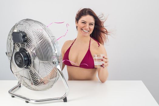 Red-haired smiling woman in a bikini drinks a cold drink and enjoys the blowing wind from an electric fan on a white background. Climate control on a hot summer day.