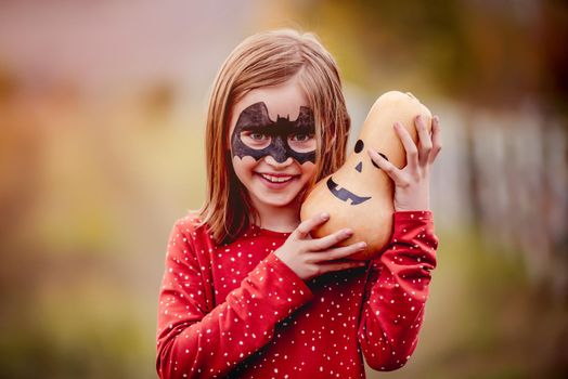halloween portrait of smiling little girl with bat mask makeup holding big pumpkin above head outdoors