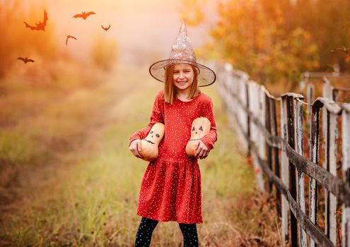 Little girl in witch hat holding halloween pumpkins standing on nature near flying bats