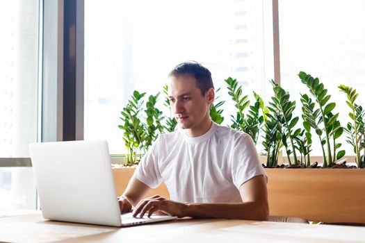 Young businessman holding mug of coffee while working on laptop computer in store with healthy food, male freelancer writing text on laptop keyboard while enjoying cup of cappuccino in modern cafe