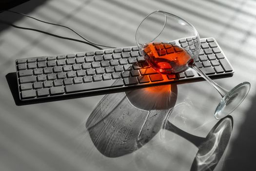 A glass of red wine lies on the keyboard on a white table with a shade from the blinds.