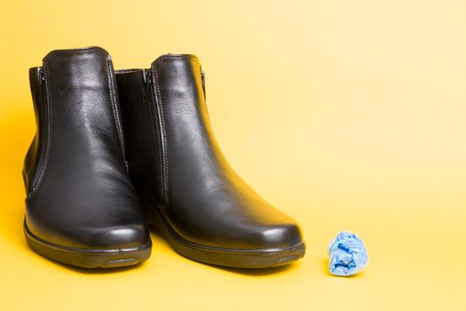 black leather boots and blue disposable shoe covers on a yellow background copy space