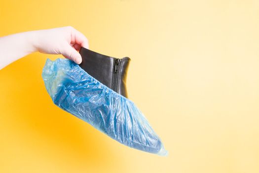 female hand holds a black leather female boot in boot covers, yellow background