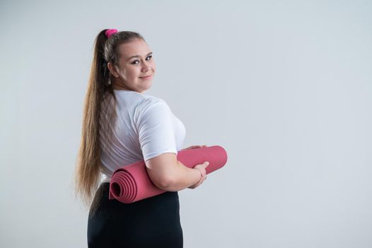 Young fat caucasian woman holding a sport mat. Charming plus size model in sportswear stands on a white background.