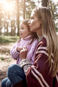 Young stylish woman is holding her daughter in forest. Woman is wearing a stylish hat and knitted coat, girl is in pink bright sweater