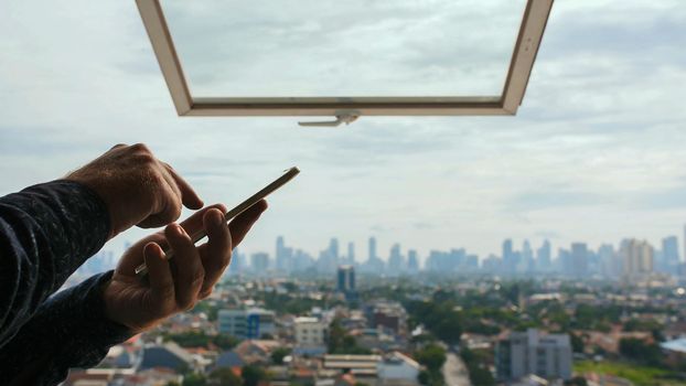 A man types a message on a smartphone against the background of an open window and a panorama of the city of Jakarta. Hands close up