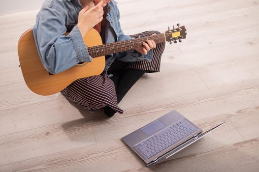 Young hipster woman sitting on the floor at home and watching guitar tutorials on laptop. Distance learning music in quarantine.