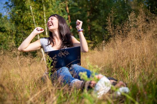 happy young woman with laptop on a sunny lawn. freelancer works in nature. student studies remotely on nature landscape outdoor