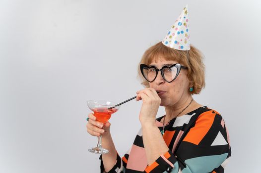 Portrait of a smiling elderly woman in a festive cap and glasses with hearts drinking a cocktail on a white background.