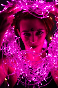 Woman with witch makeup and an earring in her nose. The girl holds a garland of flickering pink lights near her face. Light bulbs illuminate the witch's face in the dark