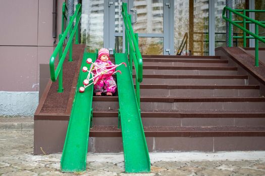 cute toddler drags a toy stroller along the ramp of the stairs. Slope walkway for disabled people, people pushing strollers, carts with stainless bars to prevent falling