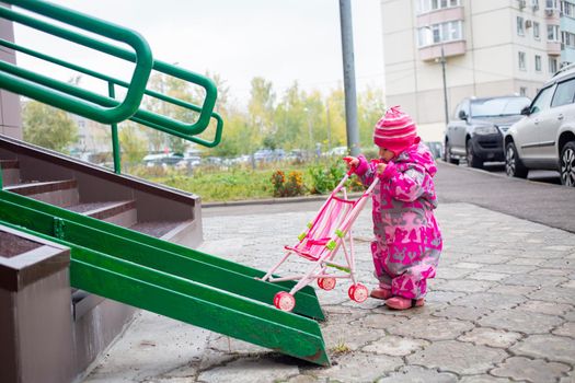 cute toddler with a toy stroller walks along steel railing ramp for wheelchair, carts and strollers. gentle descent from the stairs outdoors