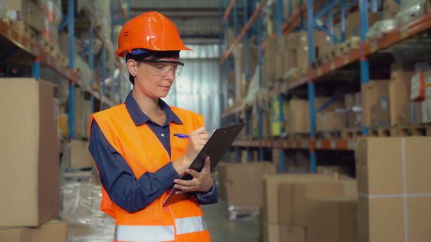 Manager female working at warehouse. Attractive young woman worker wearing uniform hard hat and orange vest, counting box for delivery filling up form holding pen.