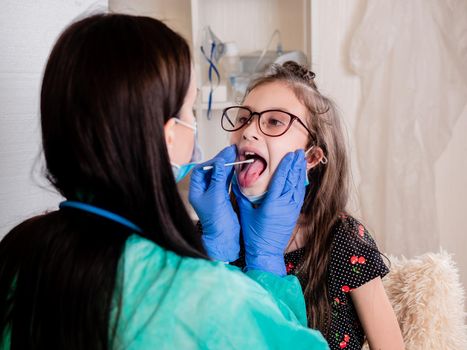 A female doctor in the office takes a test for a coronavirus infection from the mouth of a little girl.