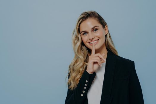 Smiling blonde girl in black coat keeping good news as secret, decides its better to wait before telling it or asking to keep silence while standing isolated on blue background. Secrecy concept