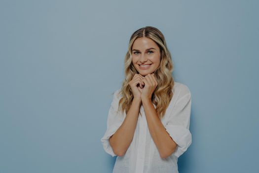 Cute young blonde woman with wavy perfect hairstyle folded hands under her chin and waiting for desired gift in excitement, dressed in white shirt. Happy facial expression and positive emotions