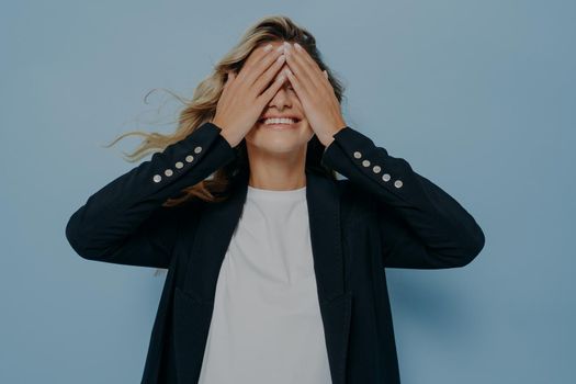 Funny blond beautiful female closing eyes with her hands, having fun and playing hide and seek while standing in stylish black blazer and white shirt. Emotional and happy mood