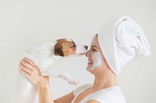 A woman with a towel on her hair and a clay mask is holding a dog. Jack Russell Terrier licks the mask from the owner face