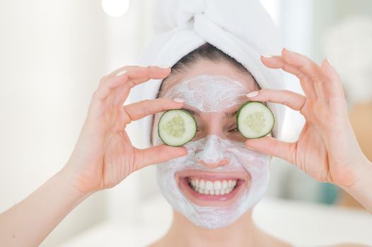 Cheerful woman with a towel on her hair and in a clay face mask and cucumbers on her eyes. Taking care of beauty at home.
