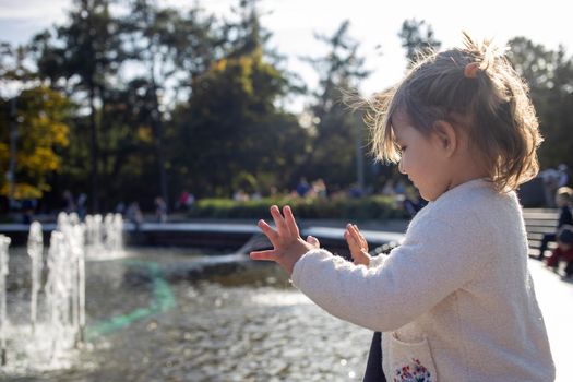 adorable little girl looks at the fountain. toddler in a park with fountains on a sunny day.. family weekendю little girl looking into a fountain in the park