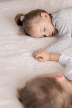 Childhood, sleep, relaxation, family, lifestyle concept - two young children 2 and 3 years old dressed in white and beige bodysuit sleep on a beige and white bed at lunch holding hands top view