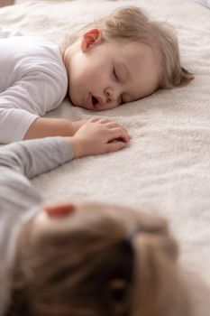 Childhood, sleep, relaxation, family, lifestyle concept - two young children 2 and 3 years old dressed in white and beige bodysuit sleep on a beige and white bed at lunch holding hands top view