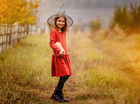 Little girl in witch hat holding candle and pumpkin standing on nature near flying bats